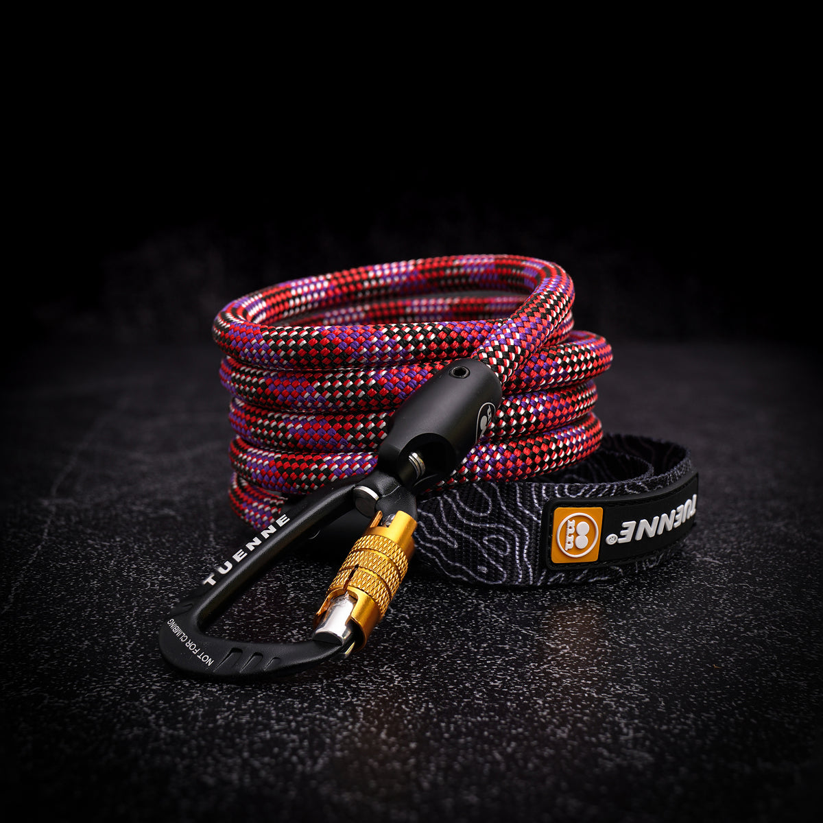 A coiled climbing rope with a red, black, and white patterned braided design, attached to a black carabiner with a gold metal lock and a black branded wrist strap on a dark textured surface.