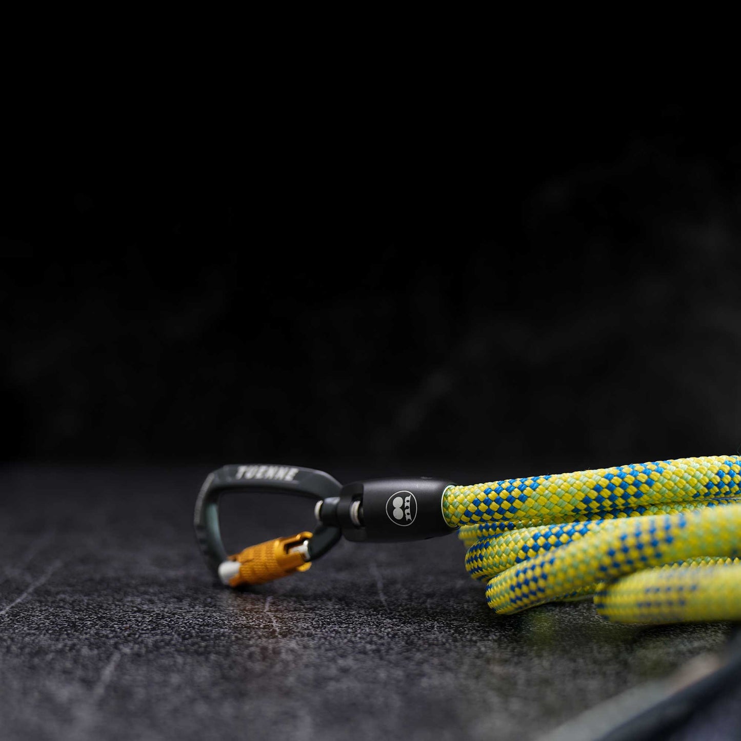 A close-up of a climbing carabiner with a bright yellow and blue patterned rope attached, photographed on a dark textured surface against a black background.