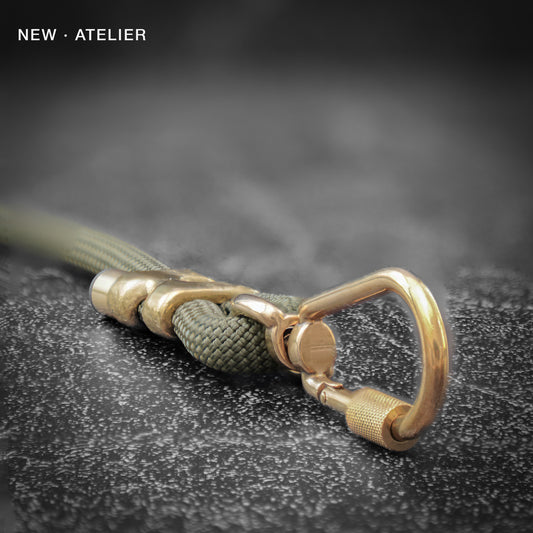 Brass clasp with green cord on a textured black surface, featuring 'NEW ATELIER' branding.