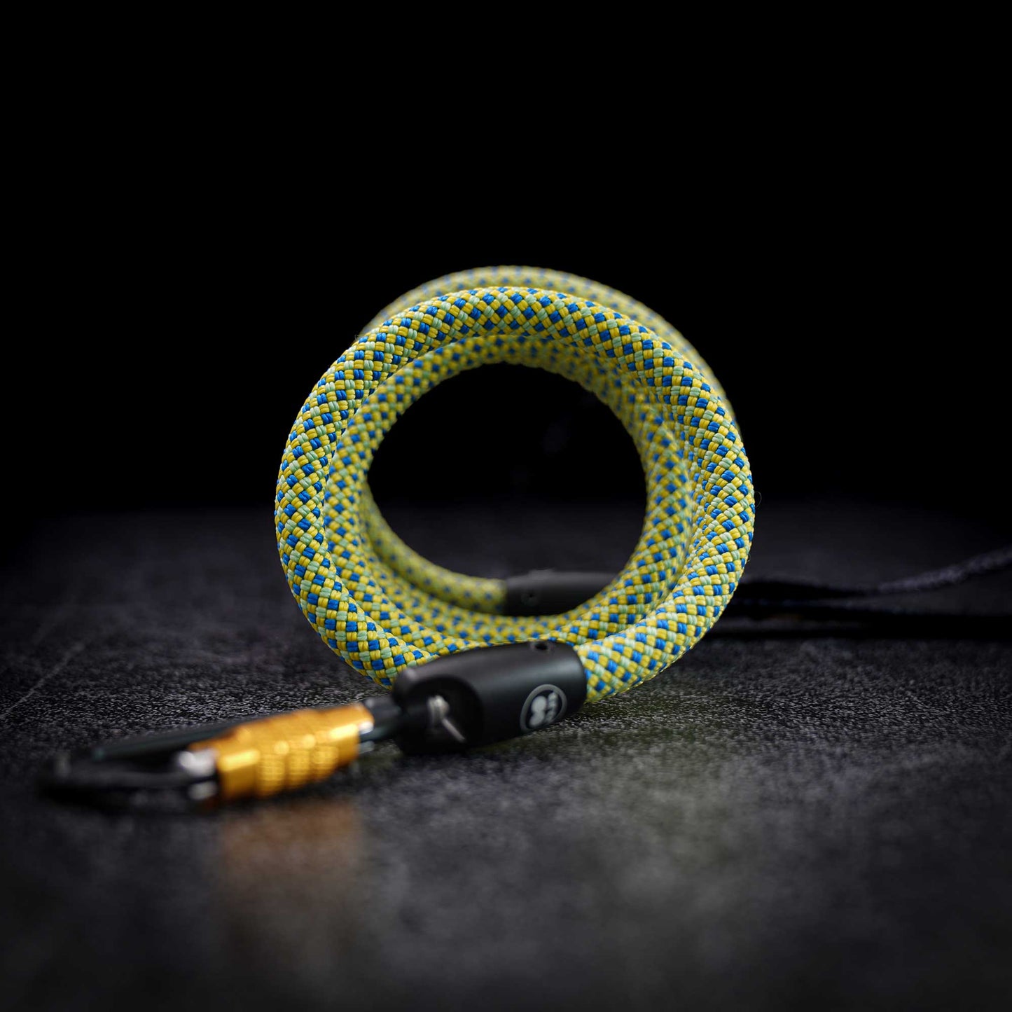 A coiled green and yellow braided rope keychain with a black plastic lock and a gold-colored carabiner, set against a dark textured background.