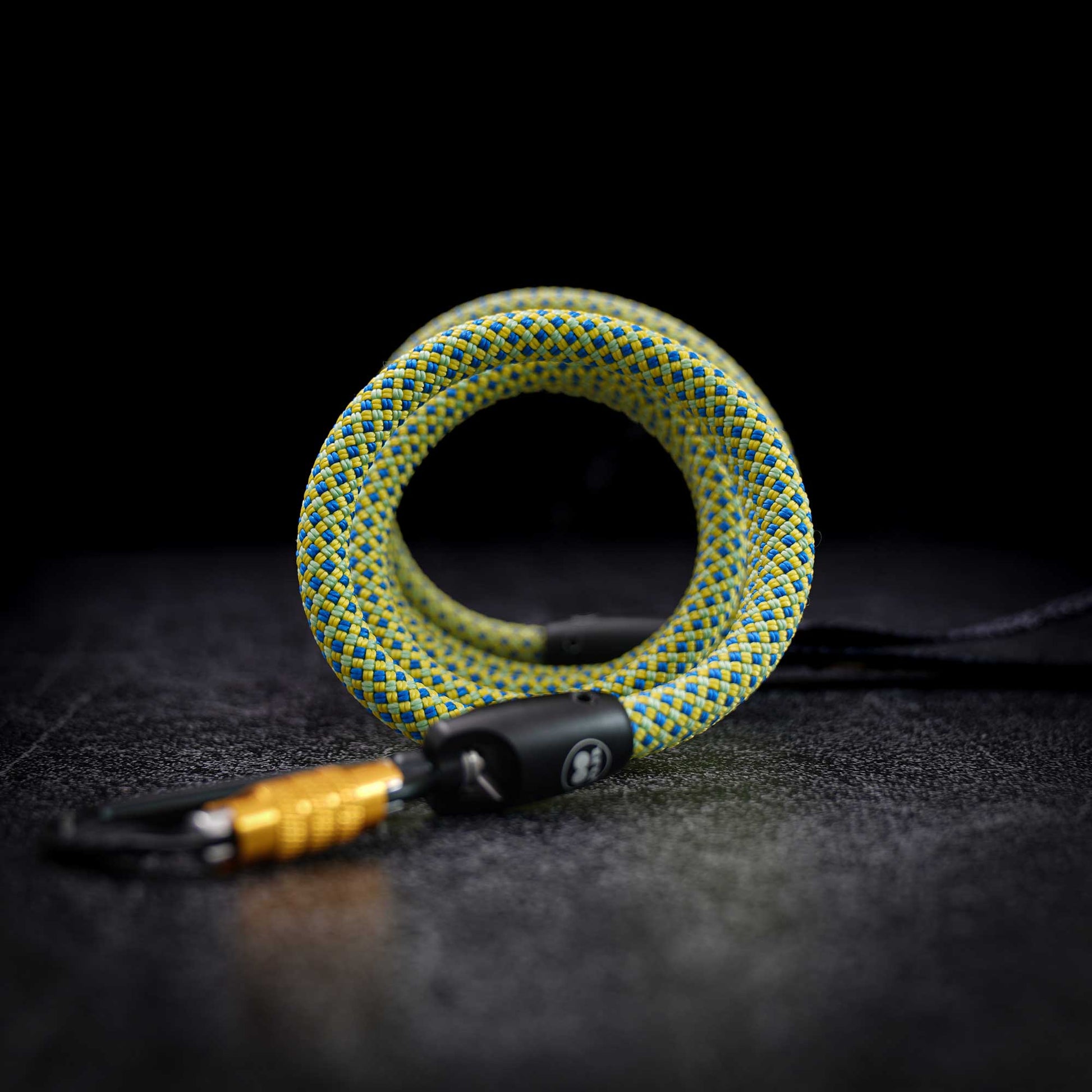 A coiled green and yellow braided rope keychain with a black plastic lock and a gold-colored carabiner, set against a dark textured background.