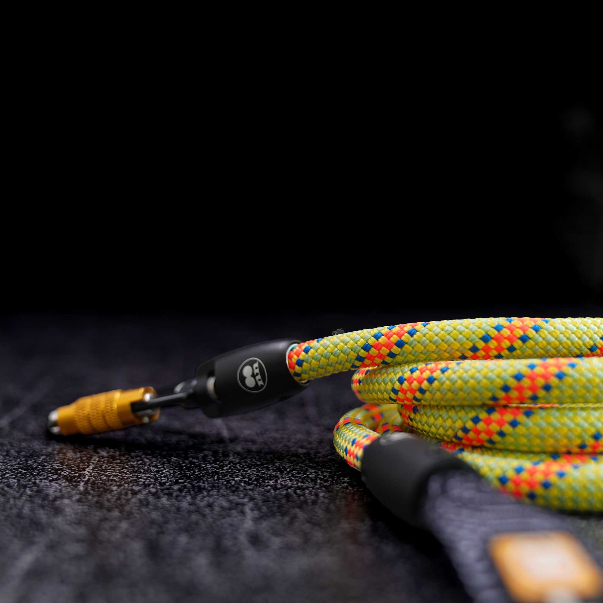A close-up of a rope dog leash with a yellow-green weave and accented with orange spiral tracer that spirals the length. the rope features a black carabiner set against a dark textured background.