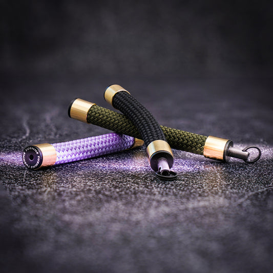 Three Atelier Rope Keychains in olive, black, and lavender, each featuring brass sleeves and a Tuenne logo-engraved endcap, resting on a textured surface.