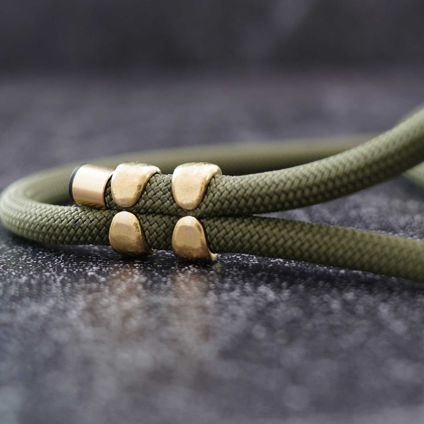 An olive green braided cord bracelet with four gold-toned metal beads, shown in a close-up shot against a dark textured background.