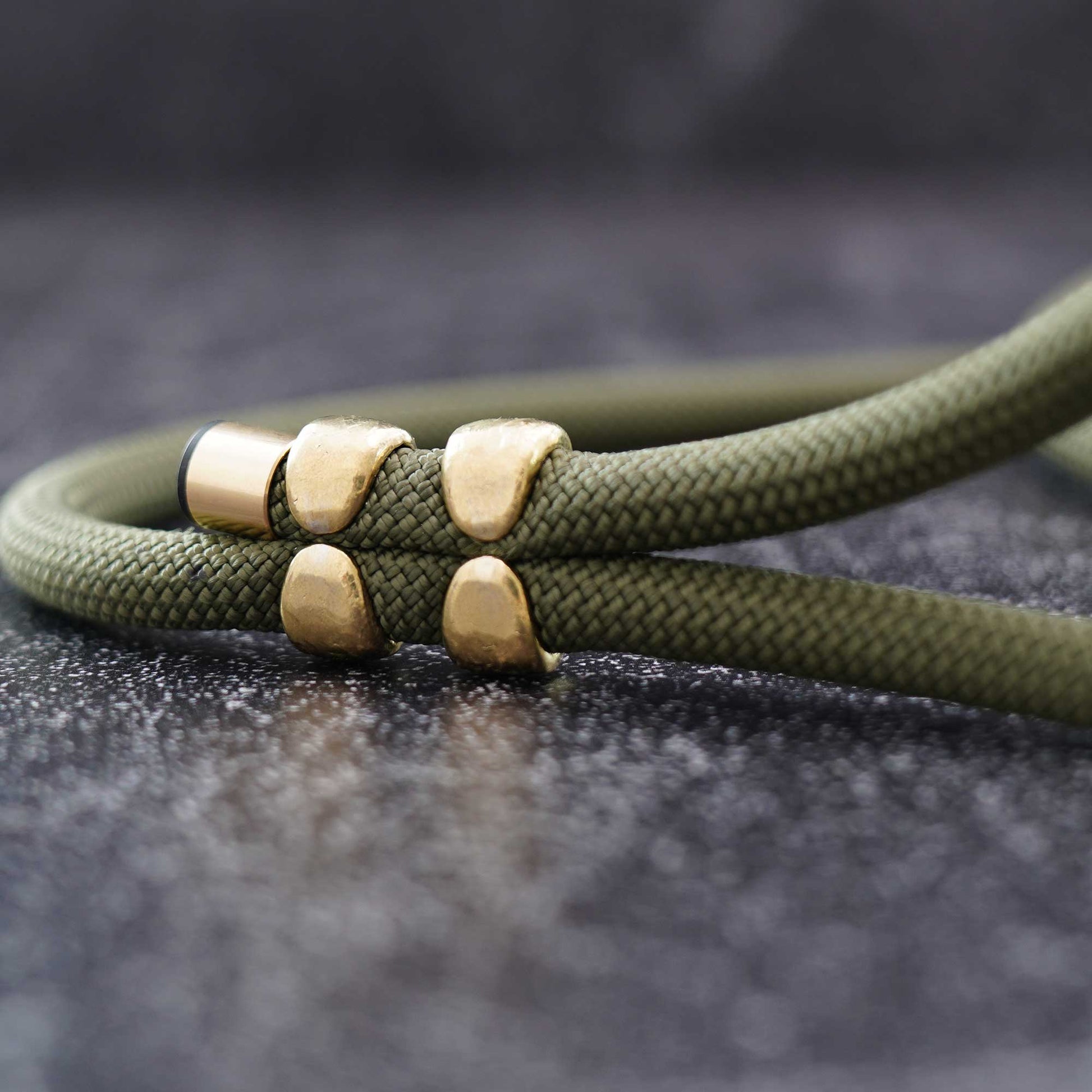 An olive green braided cord bracelet with four gold-toned metal beads, shown in a close-up shot against a dark textured background.