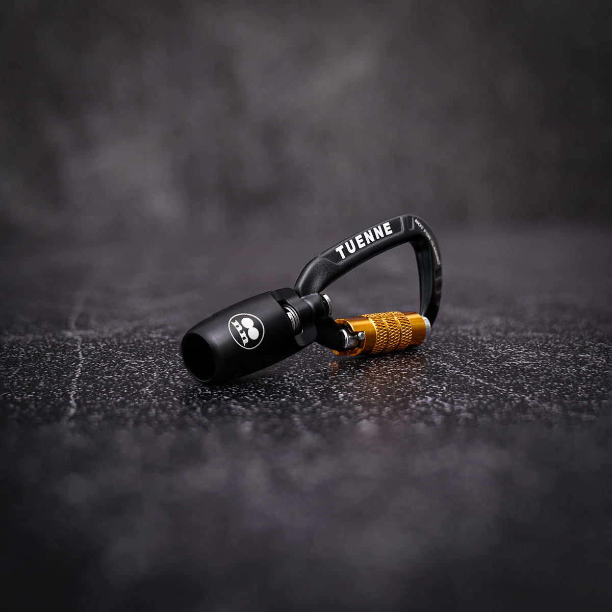 A black metal carabiner with a textured gold accent and white 'TUENNE' branding, resting on a dark textured surface.