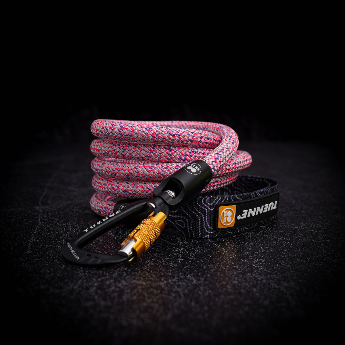 A coiled pink and blue climbing rope with a black carabiner and a black quick-release buckle, displayed on a dark textured surface. The buckle strap features the brand name 'TUENNE' in white text with an orange logo.