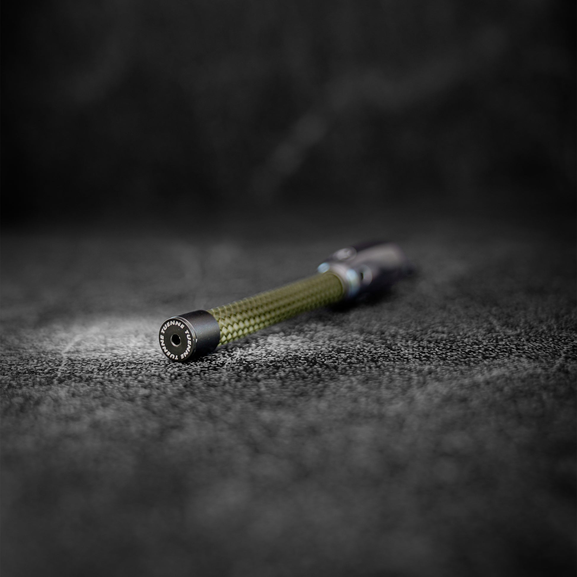 A keychain with a textured olive green climbing rope and black metal accents with a tuenne logo, resting on a dark, textured surface with a shallow depth of field.