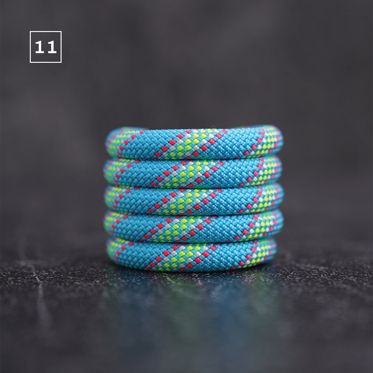 A stack of five braided rope rings in a blue, lime green, and pink pattern, displayed on a dark textured surface with a dark gray background.