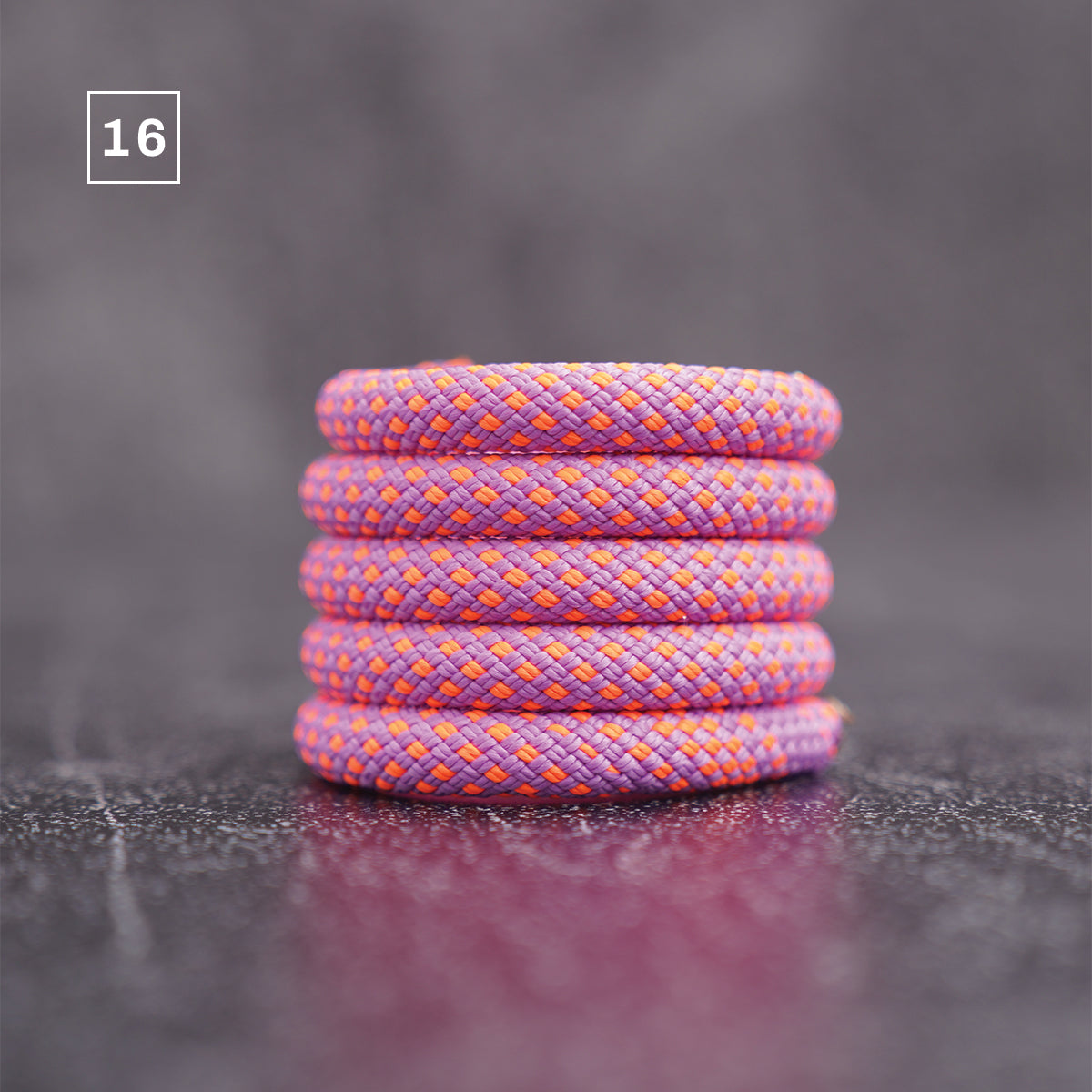 A stack of five braided elastic bands in a purple and orange pattern, set against a dark gray textured background with a '16' label in the top left corner.