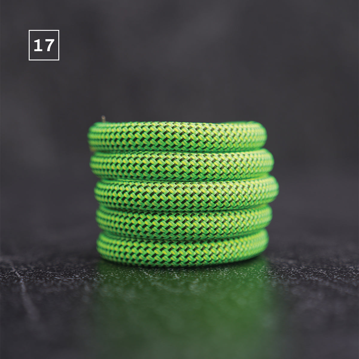 A stack of five bright neon green braided cord or rope loops, photographed on a dark textured surface with a soft reflection below. A small white square containing the number 17 appears in the upper left corner.