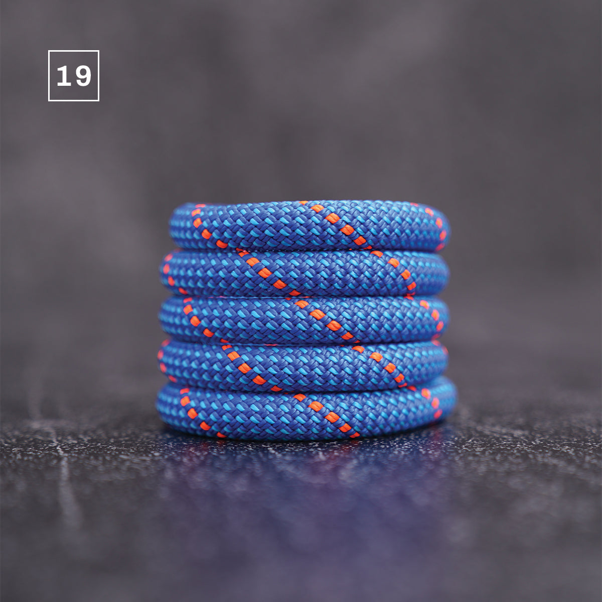 A coiled blue braided cord or rope accessory with orange stitching, photographed on a dark textured surface. A small white square with the number 19 appears in the upper left corner.