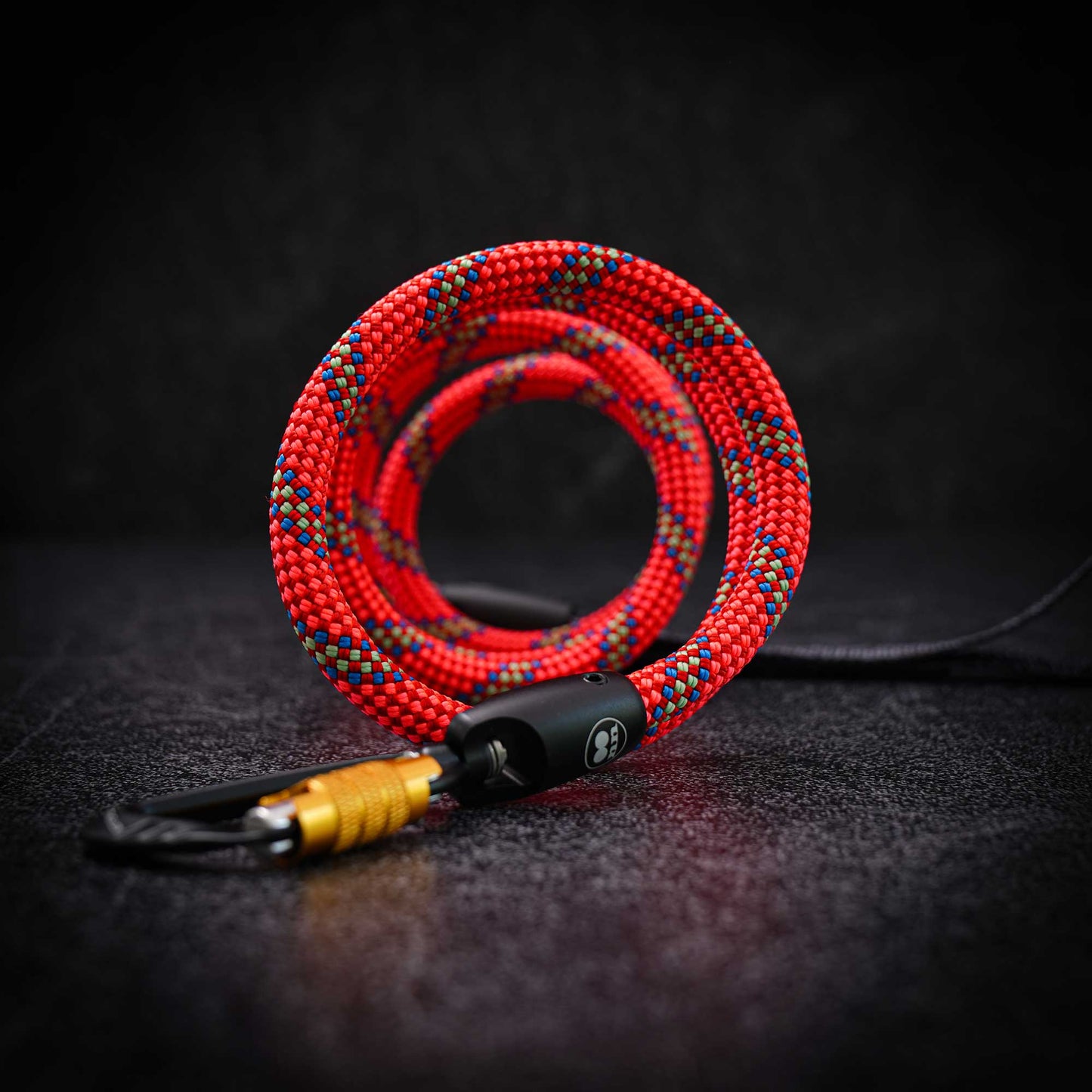 A coiled red braided rope with blue and yellow speckled pattern, attached to a black and  gold carabiner, displayed on a dark textured surface.
