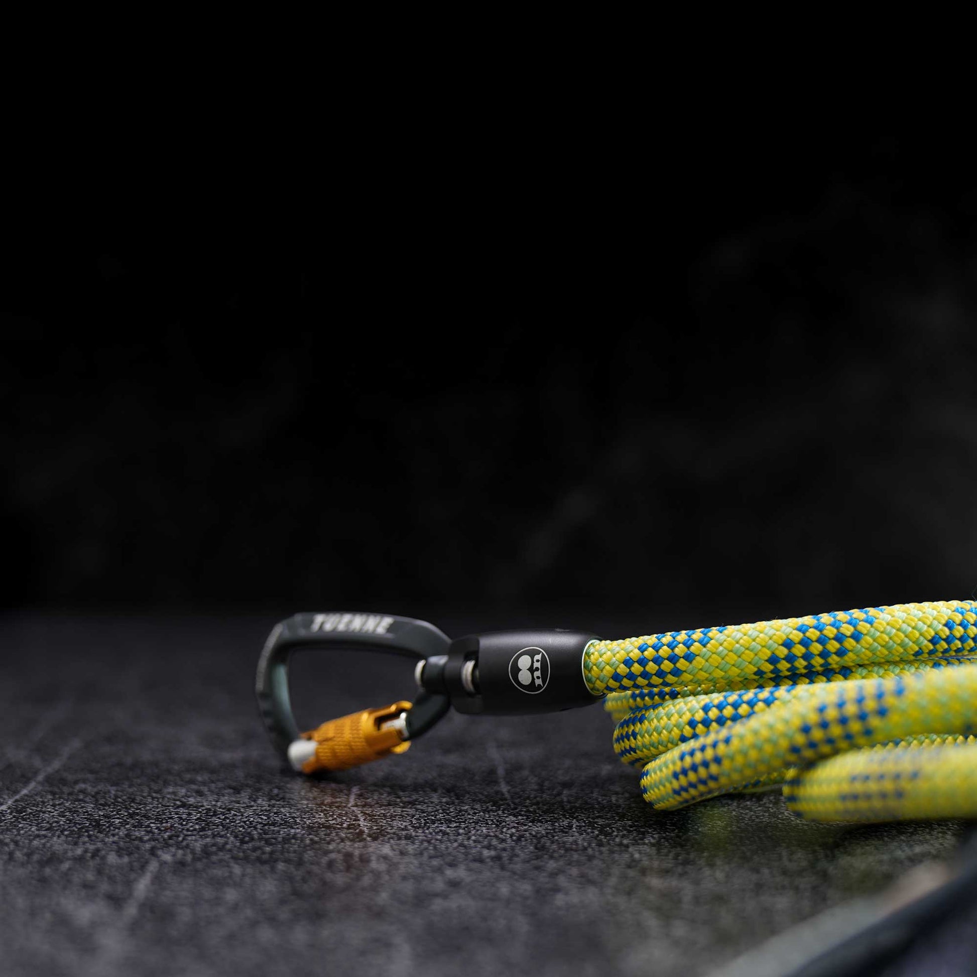 A close-up of a climbing carabiner with a bright yellow and blue patterned rope attached, photographed on a dark textured surface against a black background.