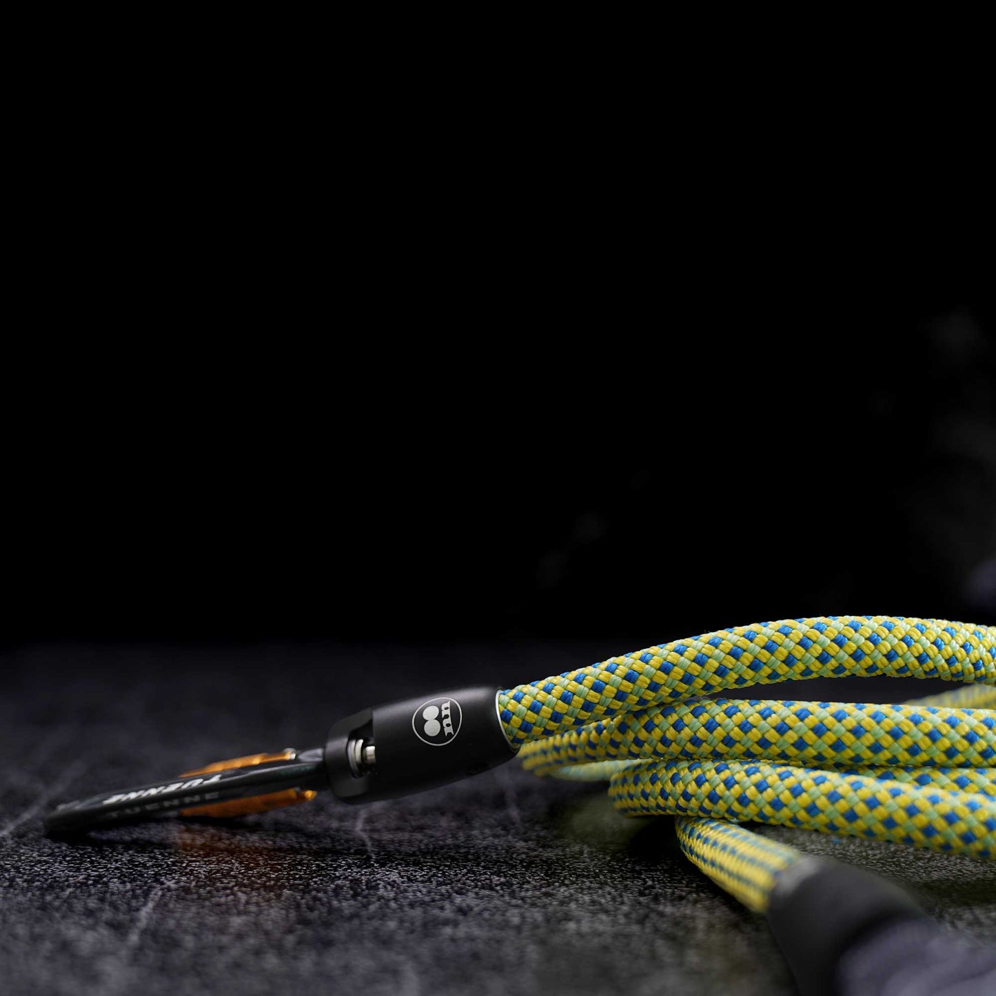 A close-up of a braided green and yellow rope with a black metal carabiner and orange accent, resting on a dark textured surface against a black background.