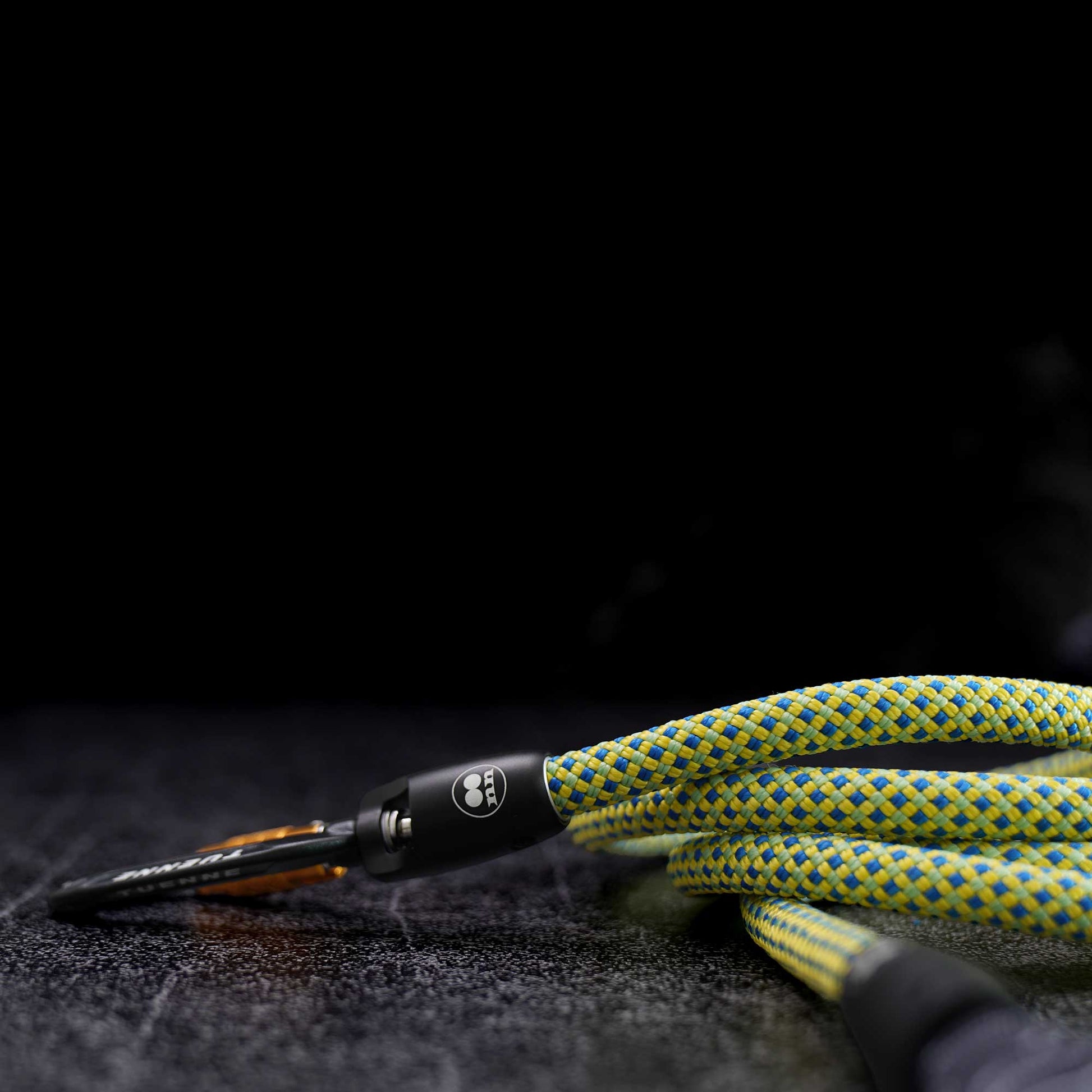 A close-up of a braided green and yellow rope with a black metal carabiner and orange accent, resting on a dark textured surface against a black background.
