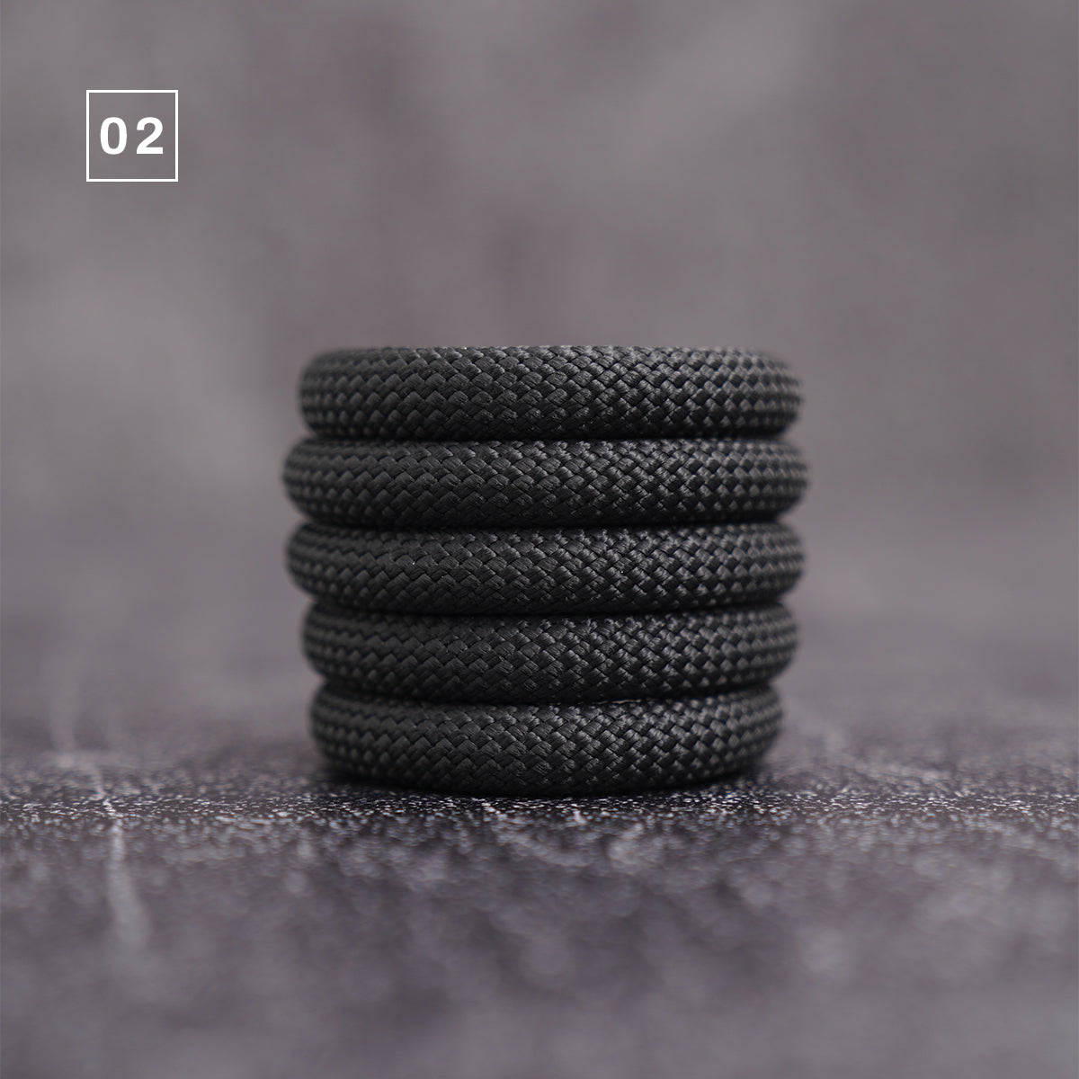 A stack of five black braided cord or rope rings, shown on a textured gray surface with a soft blurred background. A small white square in the upper left corner contains the number '02'.