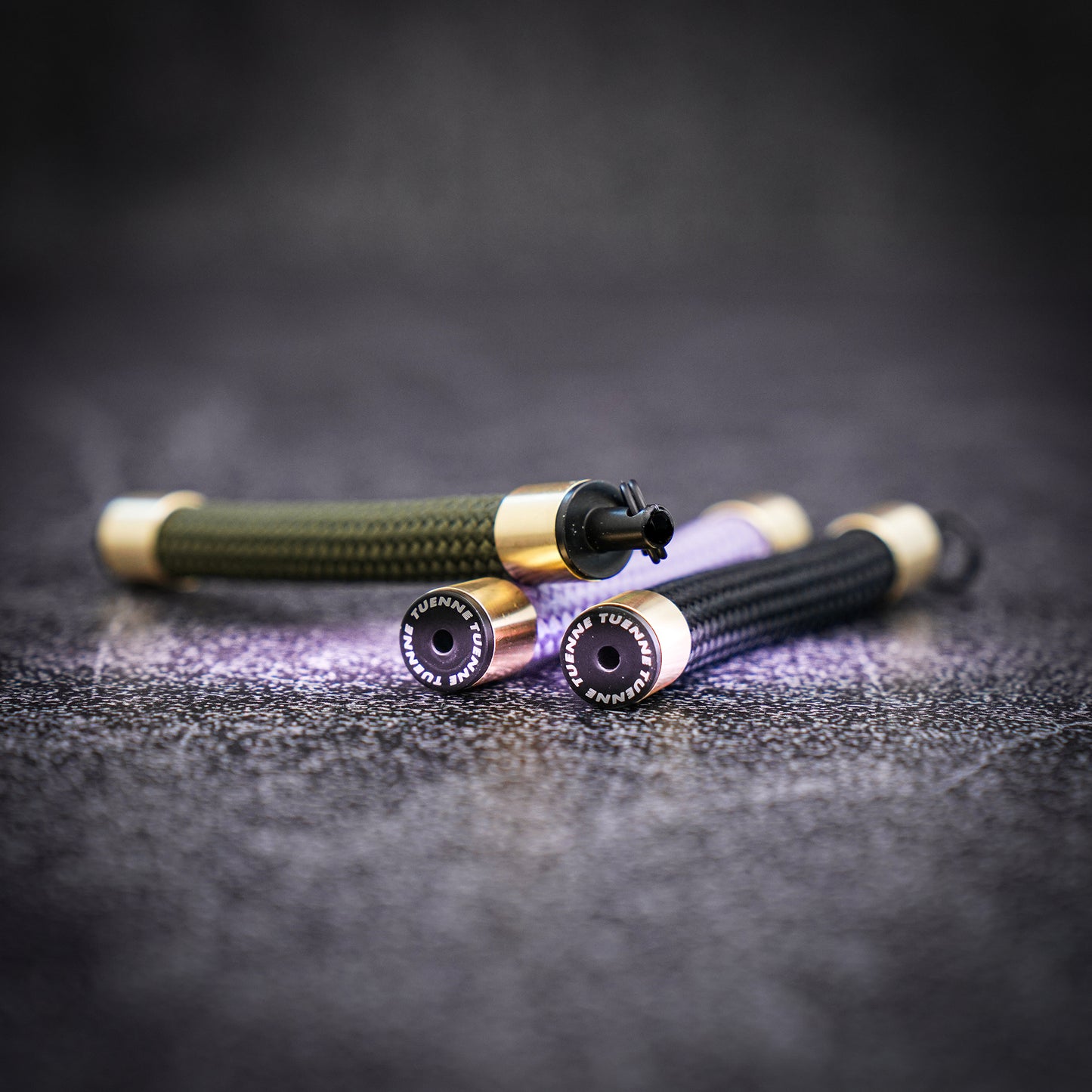 Three Atelier Rope Keychains—olive, black, and lavender—lie on a textured surface, each featuring brass sleeves and a Tuenne-branded endcap facing the camera.