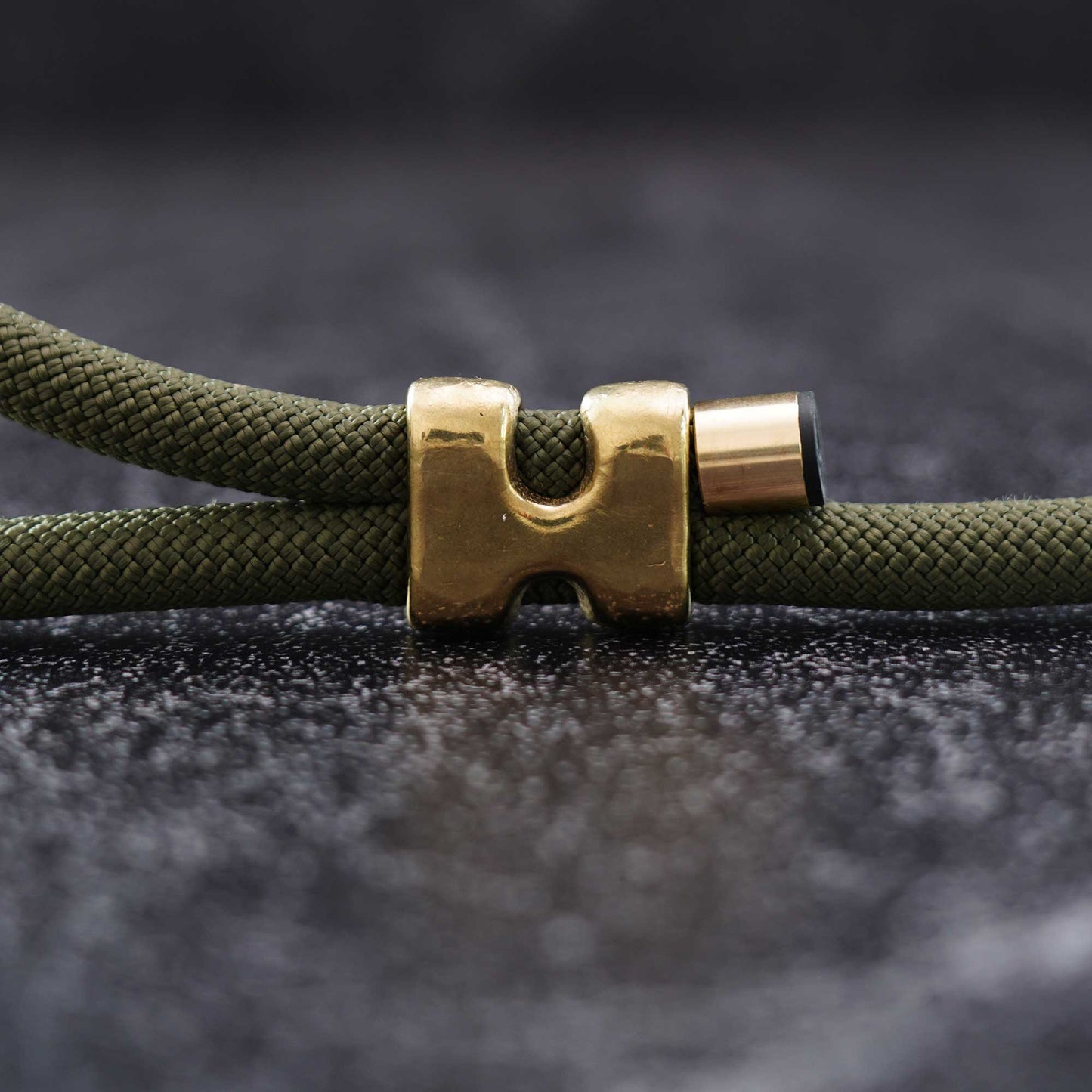 Close-up of an olive green braided cord bracelet or necklace with a polished gold-toned metal clasp on a dark textured background.