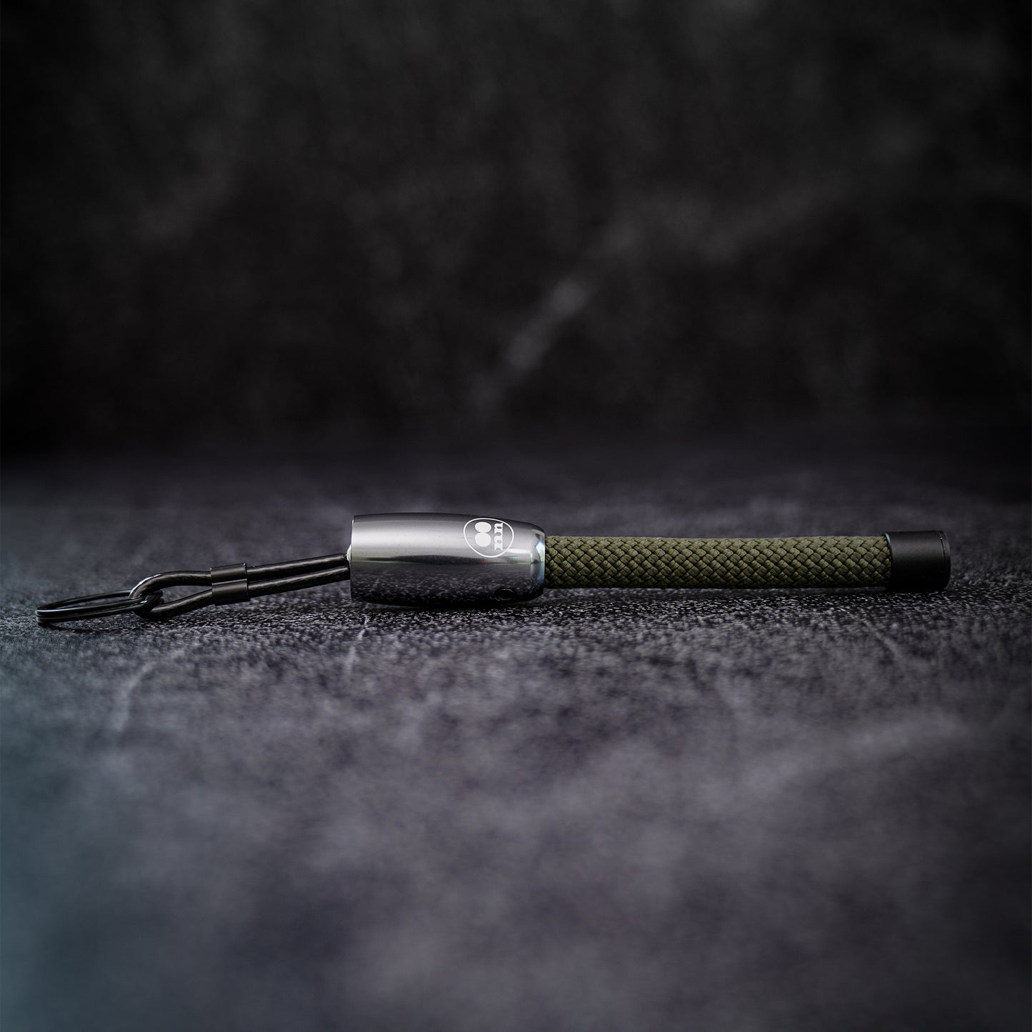 A keychain with a silver aluminum end cap, and an olive green climbing rope, resting on a dark textured surface.