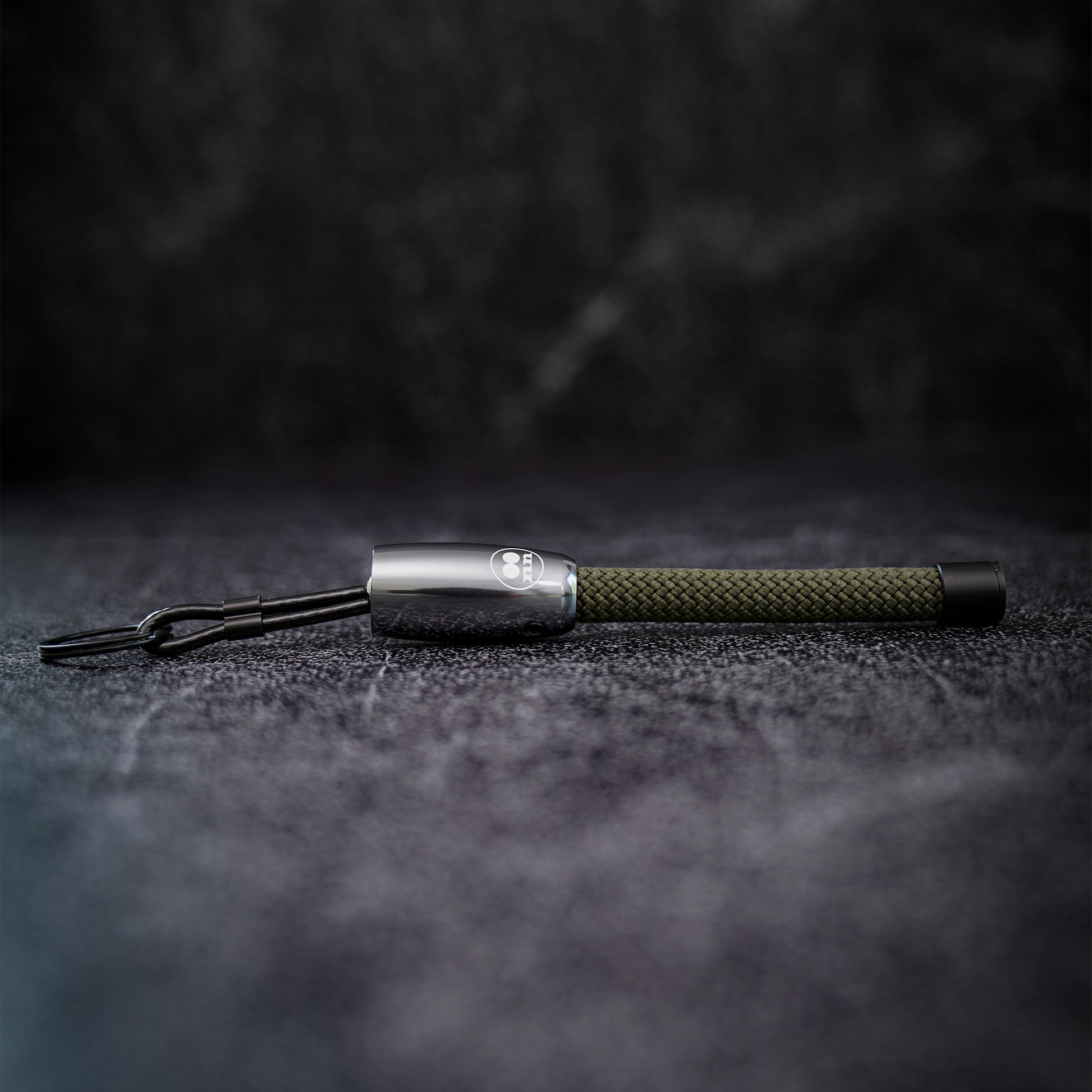 A keychain with a silver aluminum end cap, and an olive green climbing rope, resting on a dark textured surface.