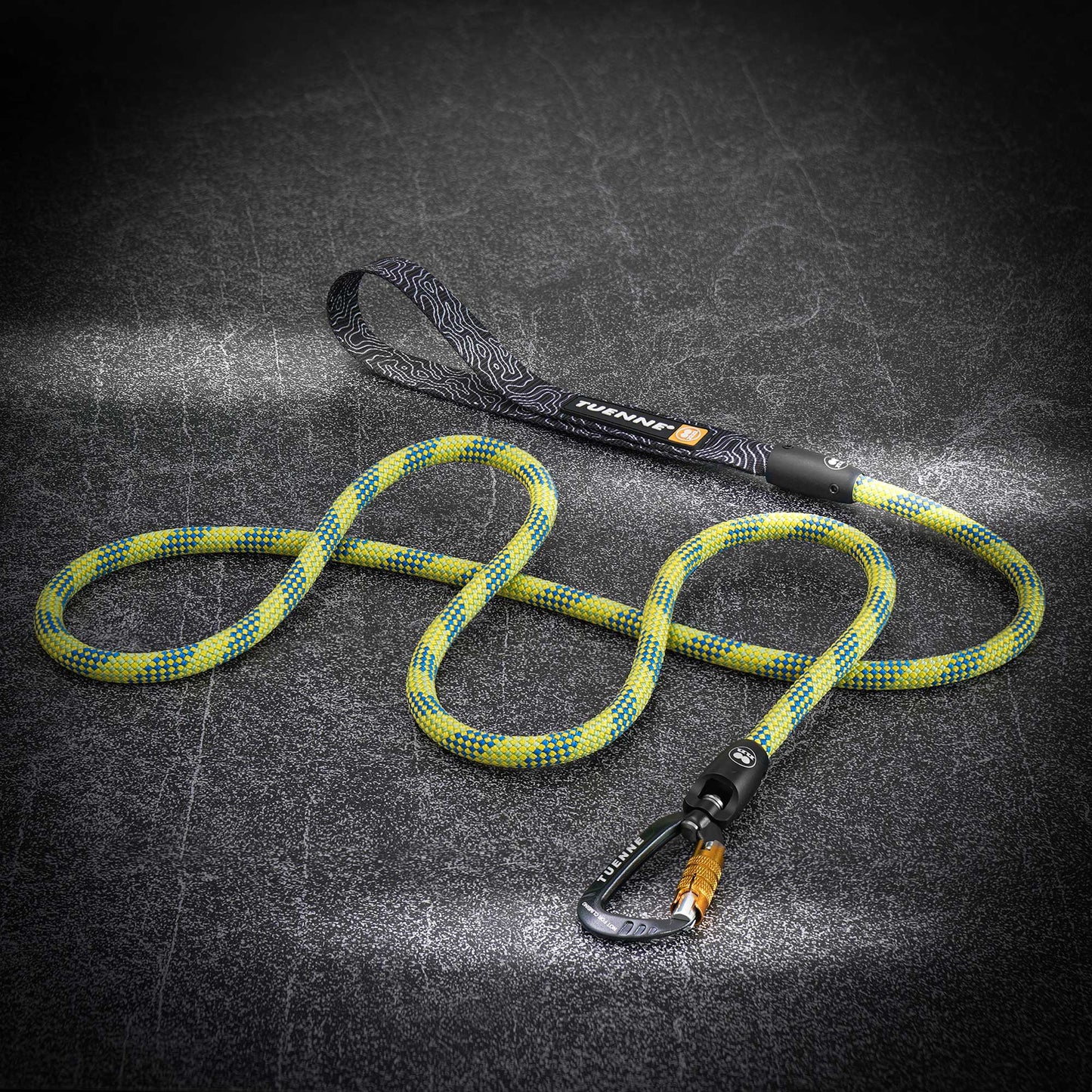 A climbing rope dog leash with a yellow and blue patterned rope, black handle, and black carabiner, displayed on a dark textured background.