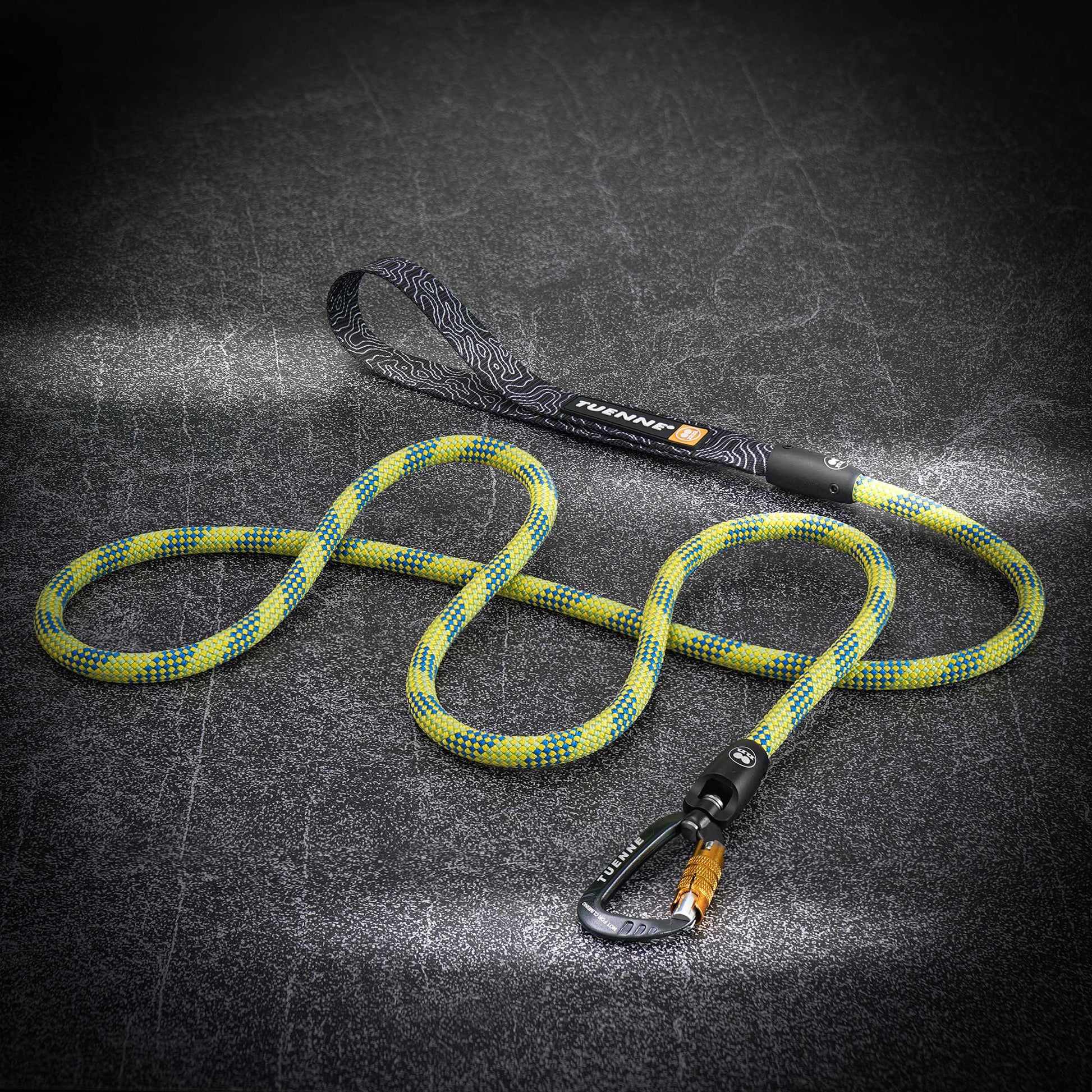 A climbing rope dog leash with a yellow and blue patterned rope, black handle, and black carabiner, displayed on a dark textured background.