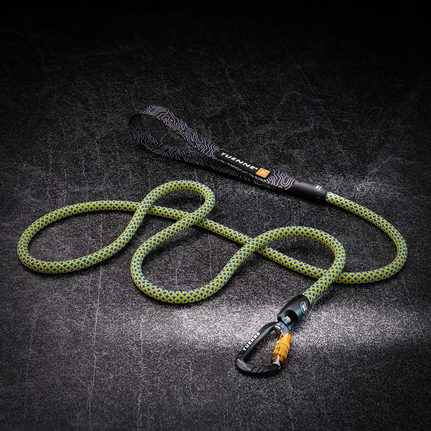 A green and yellow braided climbing rope with a black patterned wrist loop and a black carabiner with an orange accent, displayed on a dark textured background.