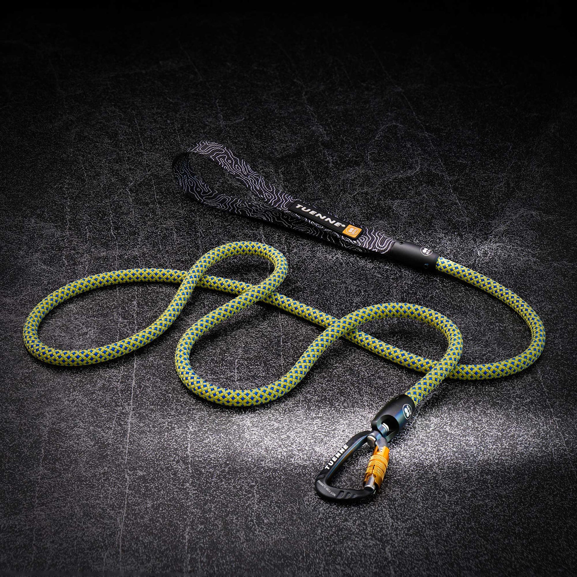 A green and yellow braided climbing rope with a black patterned wrist loop and a black carabiner with an orange accent, displayed on a dark textured background.