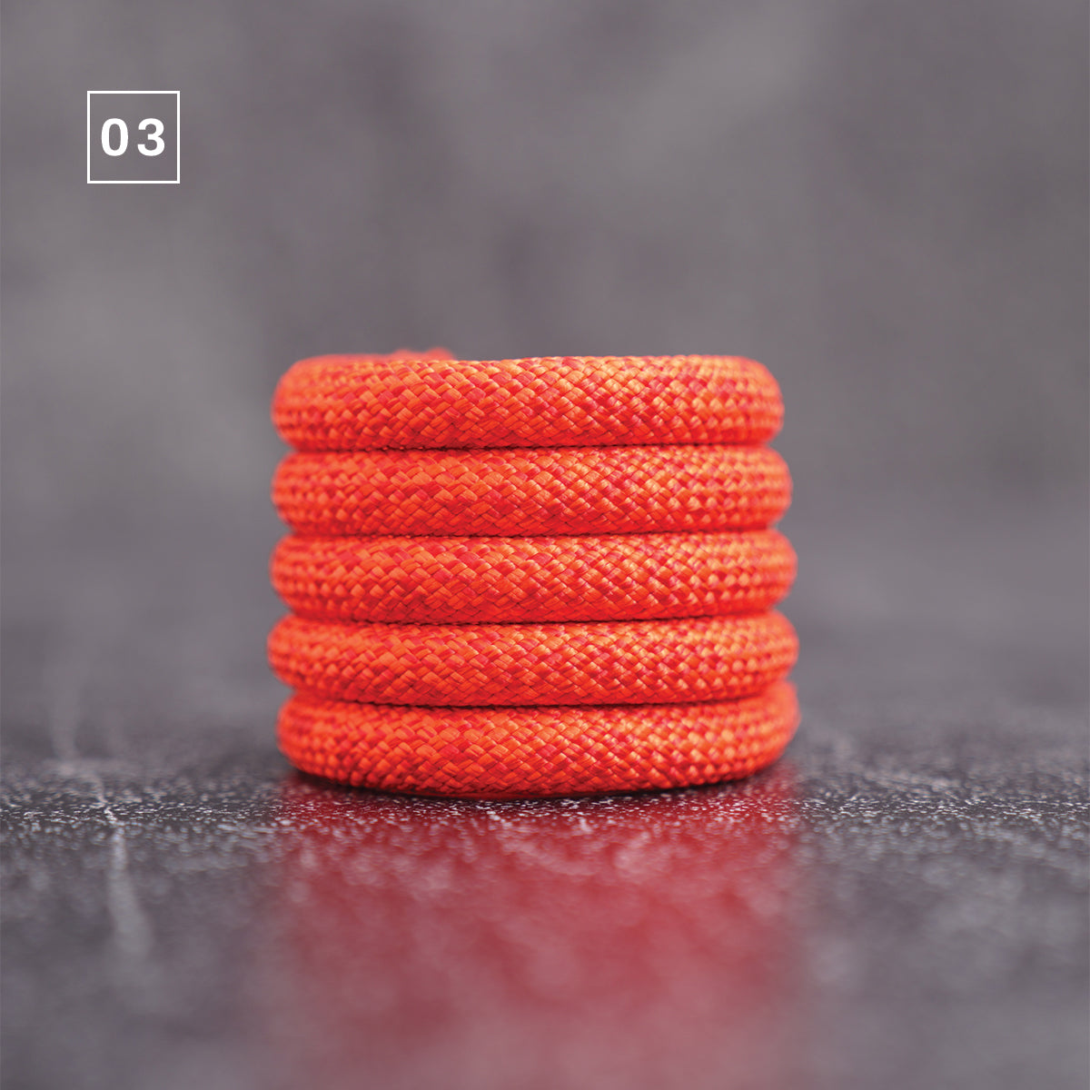 Five stacked circular orange-red braided cords or rope rings on a dark gray textured surface with a soft reflection, featuring a '03' label in the top-left corner.
