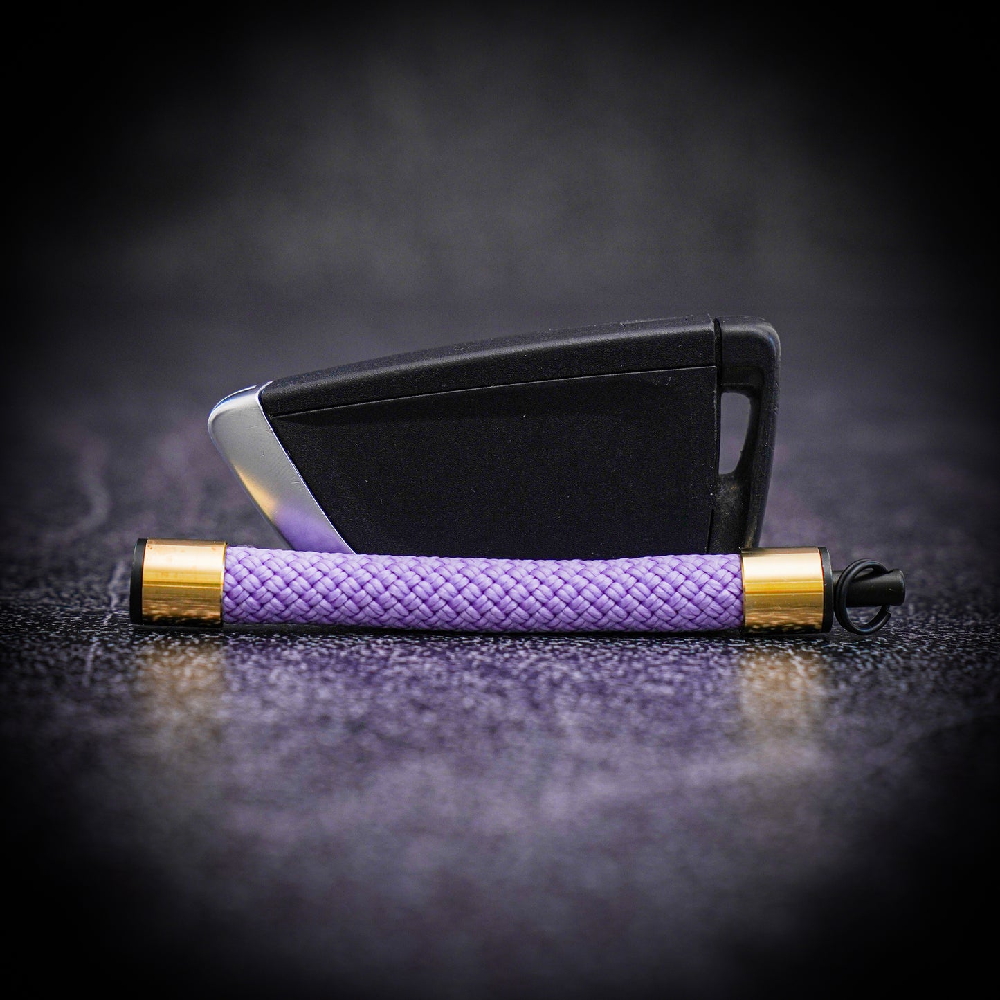 A lavender Atelier Rope Keychain with brass sleeves lies in front of a car key fob, showing its compact size and elegant design.