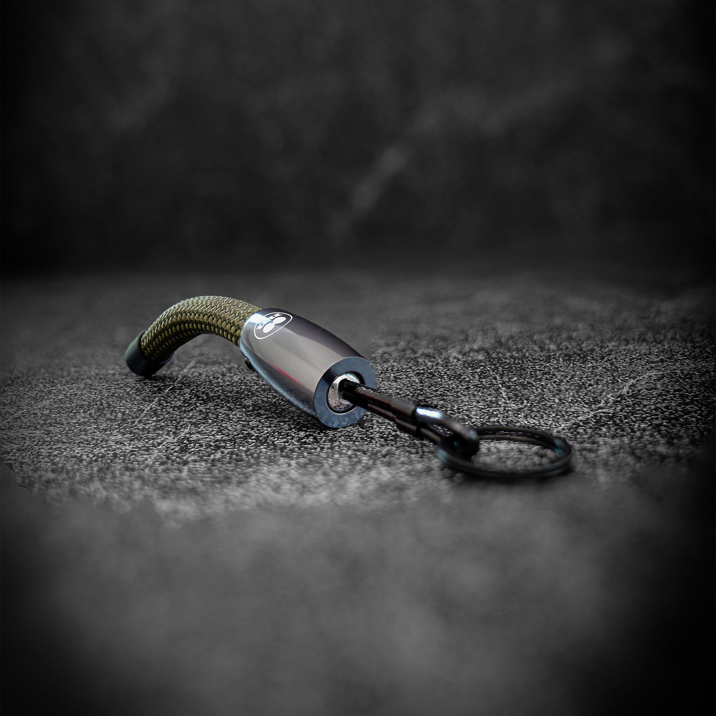 A tactical keychain with a curved olive green paracord wrap, a metallic silver cylindrical body, and a black carabiner clip attached to a black split ring, resting on a dark textured surface.