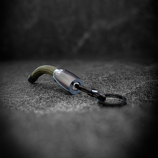 A tactical keychain with a curved olive green paracord wrap, a metallic silver cylindrical body, and a black carabiner clip attached to a black split ring, resting on a dark textured surface.