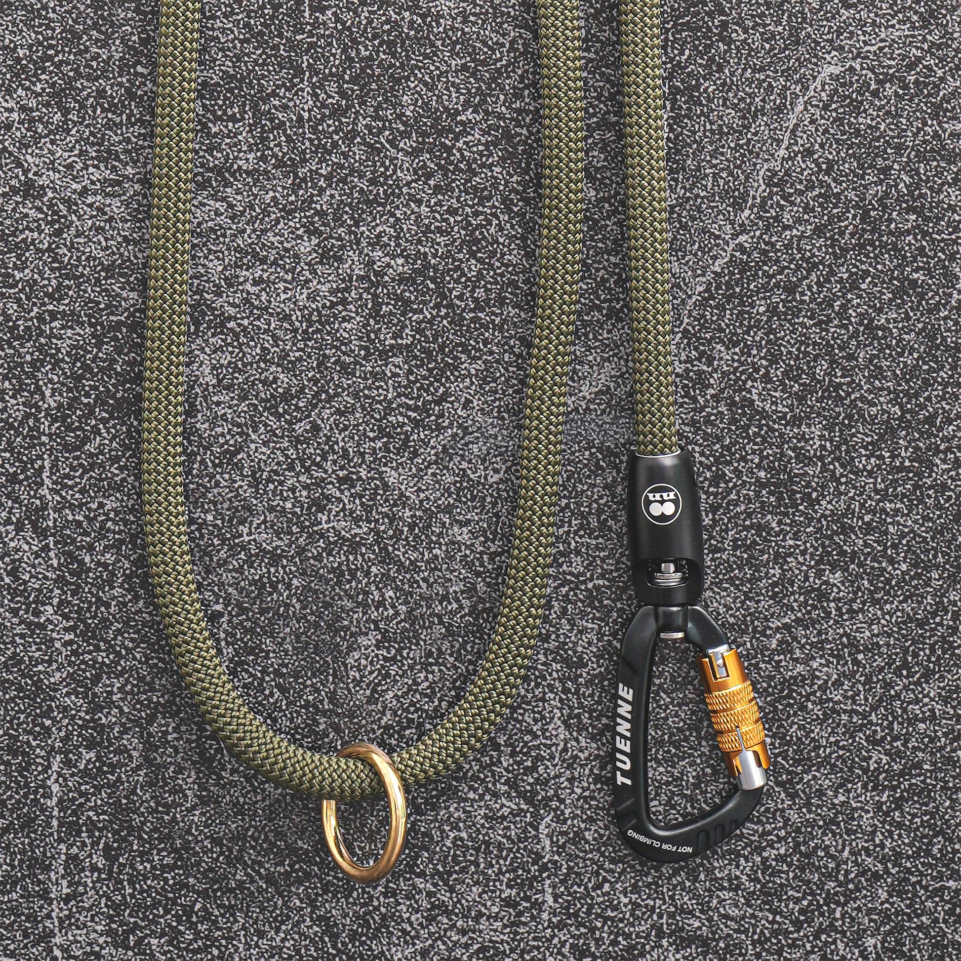An olive green paracord lanyard with a brass ring and a black carabiner featuring a yellow lock, displayed on a textured gray background.