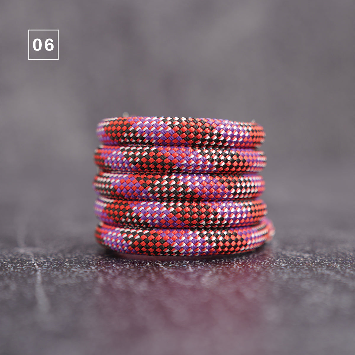 A stack of five braided elastic bands or cords in a red, purple, and black pattern, displayed on a dark textured surface with a gray background. A small white square containing the number '06' is in the top-left corner.