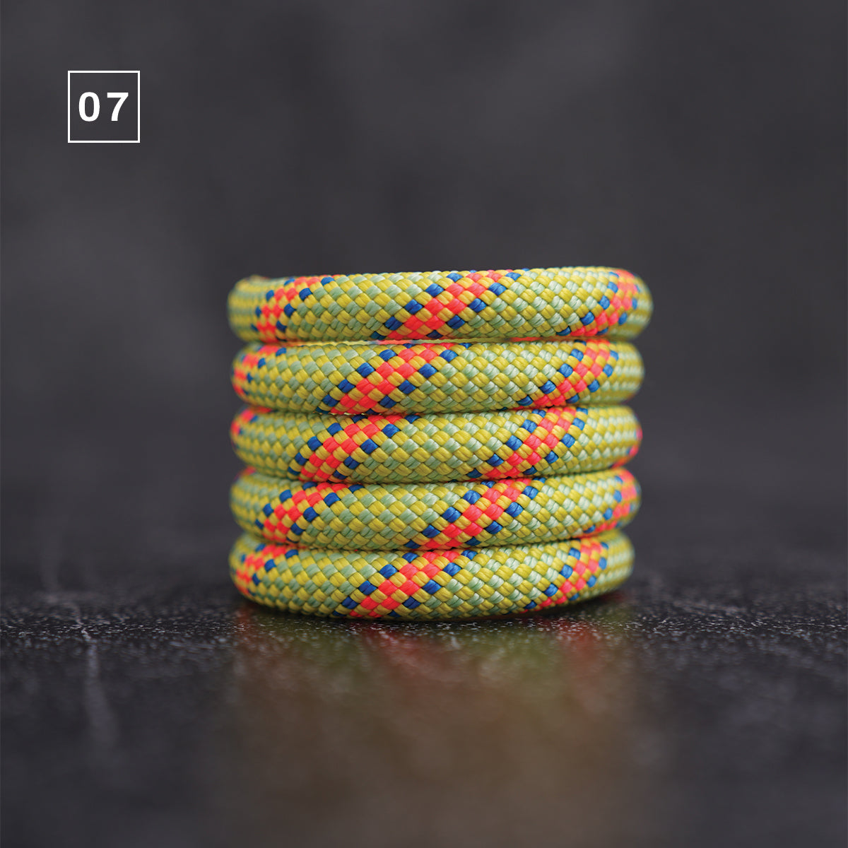 A stack of five braided rope bracelets in olive green with diagonal orange, blue, and red woven accents, displayed on a dark reflective surface with a '07' label in the top left corner.