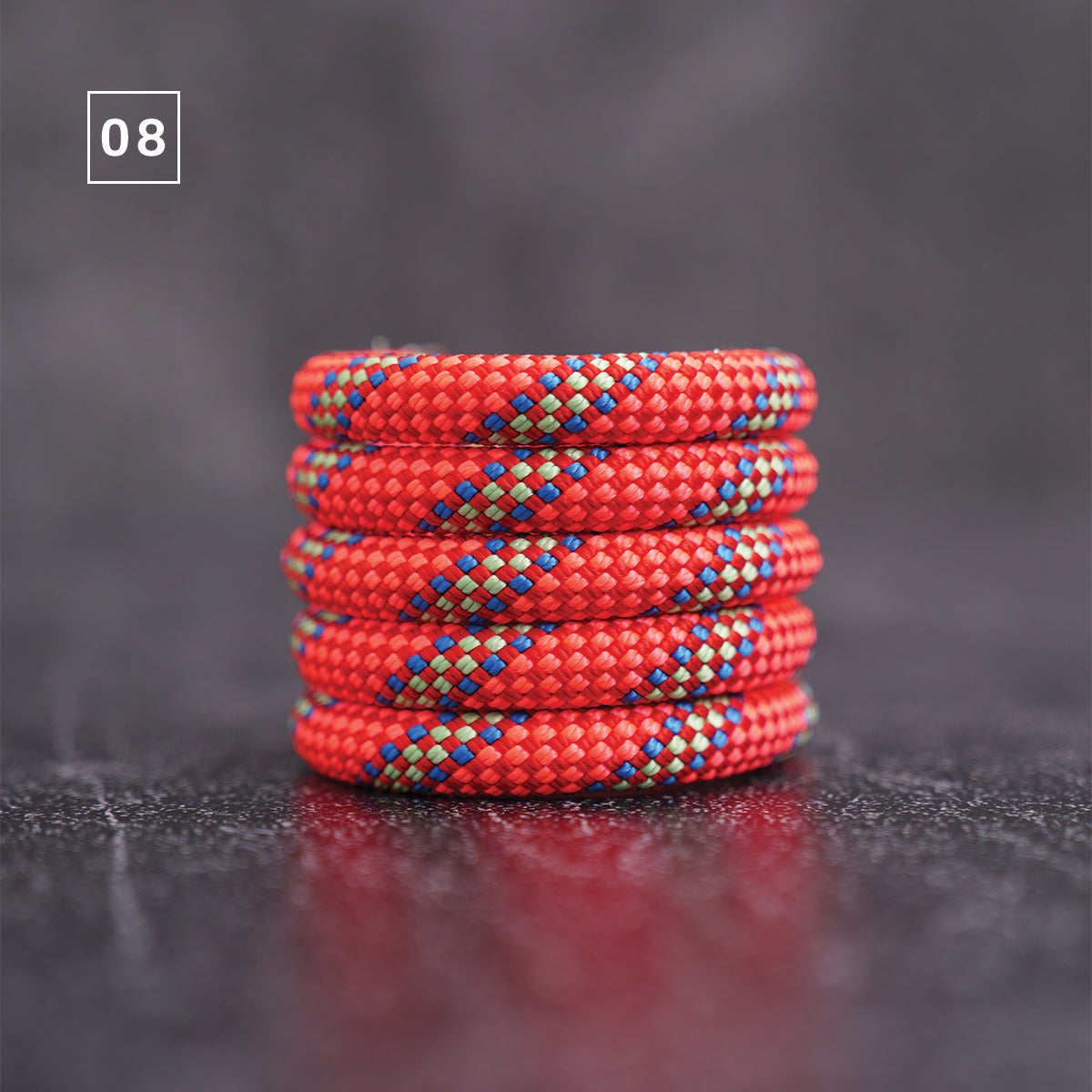 A stack of five red braided cord bracelets with blue and green woven accents, displayed on a dark textured surface with a gray background.