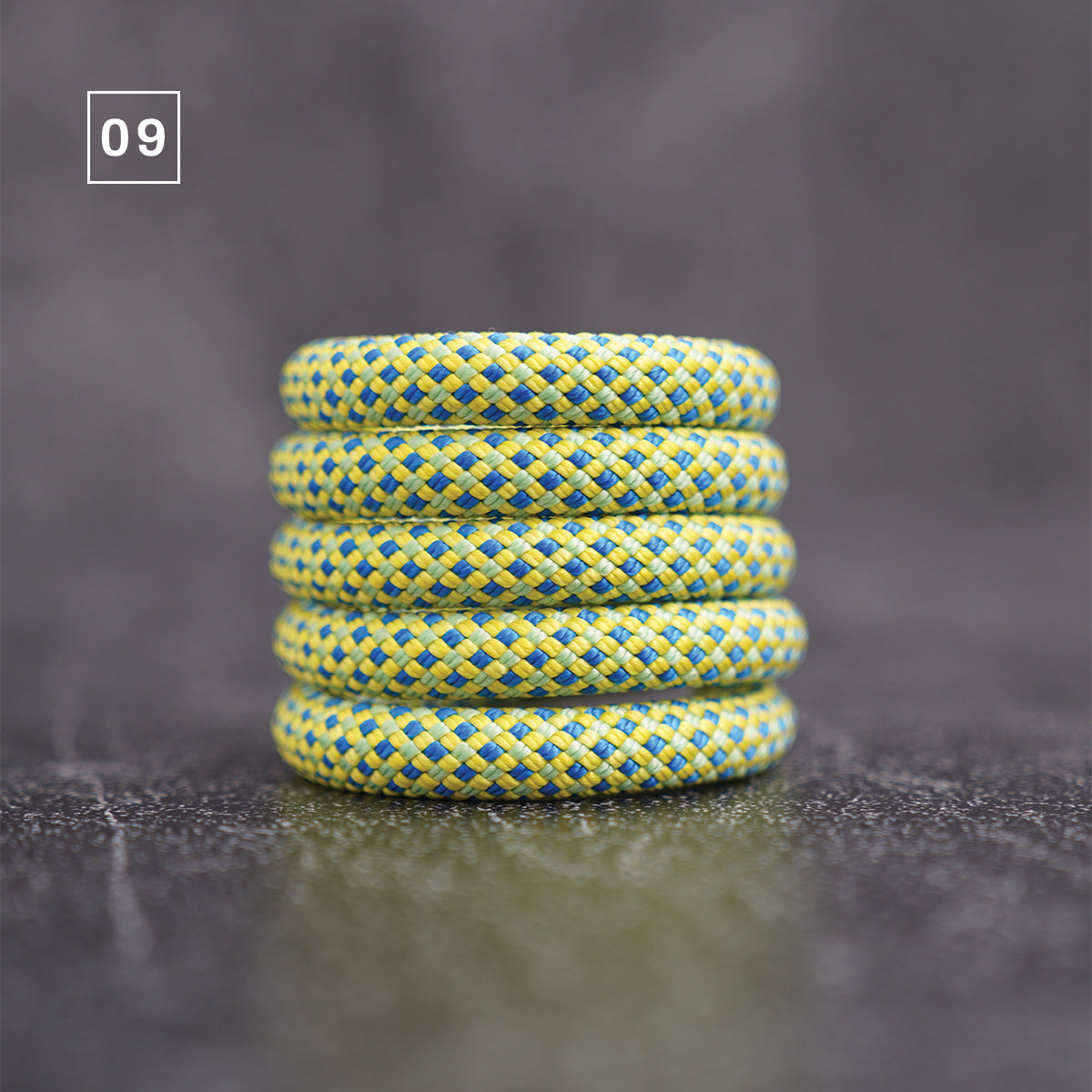 Four braided rope wristbands stacked vertically on a dark gray reflective surface. The rope has a textured pattern of yellow and blue braided strands.