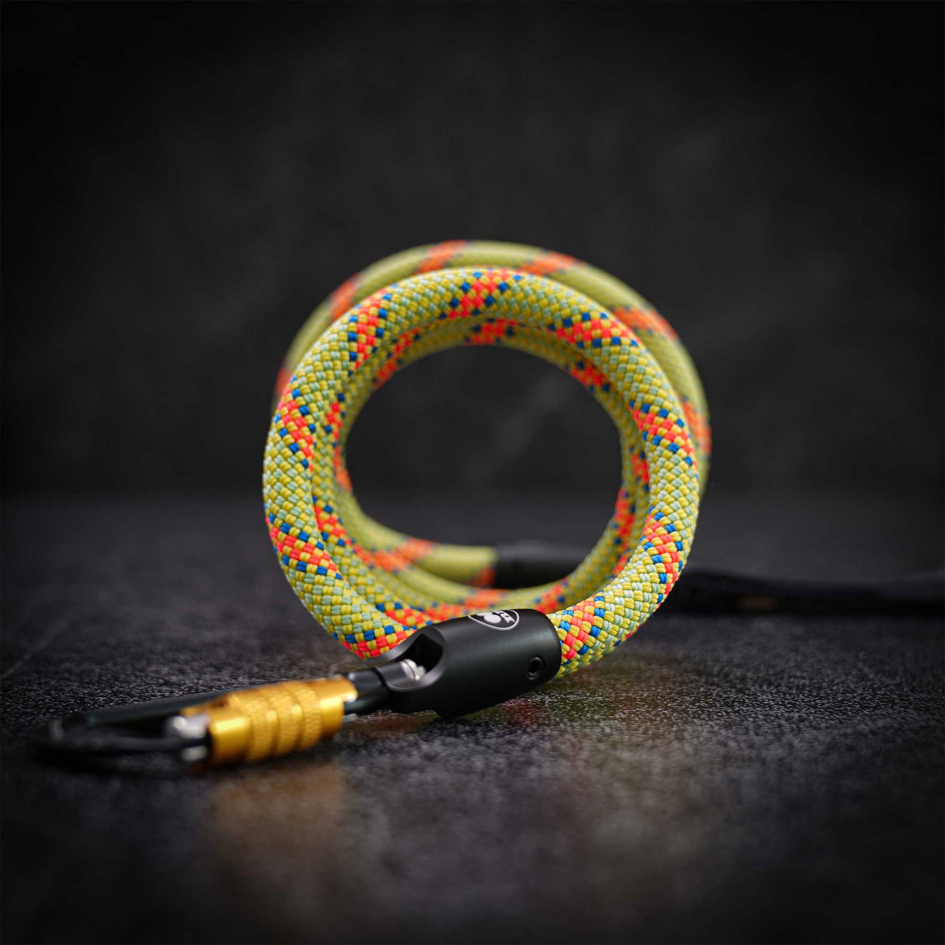 A coiled green braided rope or lanyard with orange and blue woven accents, attached to a black swivel clasp and gold-toned carabiner, photographed on a dark textured surface.