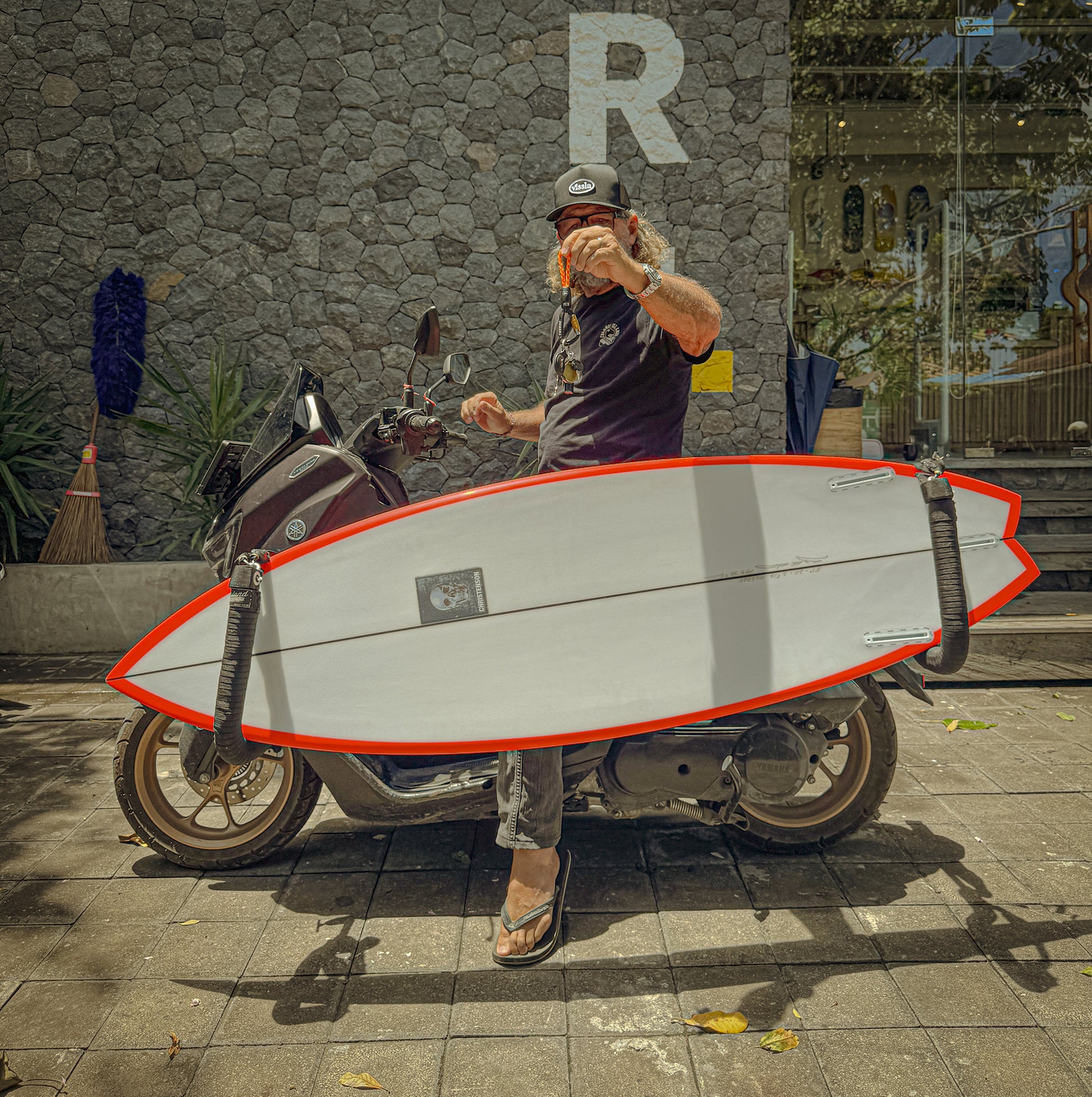 Man holding a orange-rope-keychain on a scooter with a surfboard on it. 