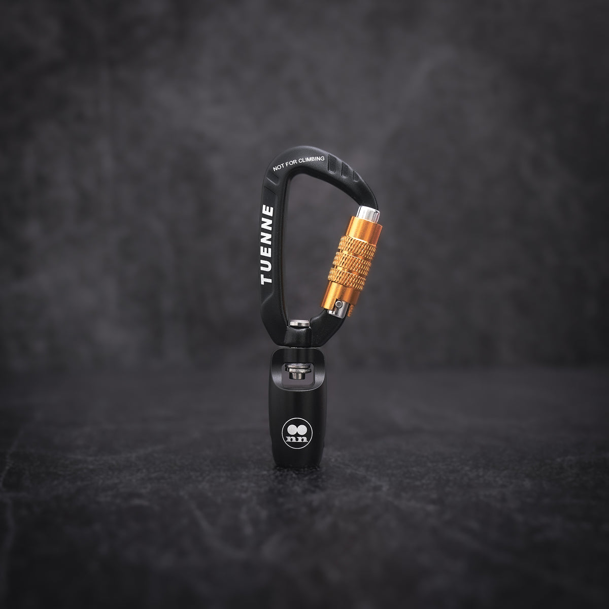 A black carabiner with a prominent textured orange-gold locking bolt, standing vertically on a dark textured surface. The brand name 'TUENNE' is printed in white on the side of the carabiner