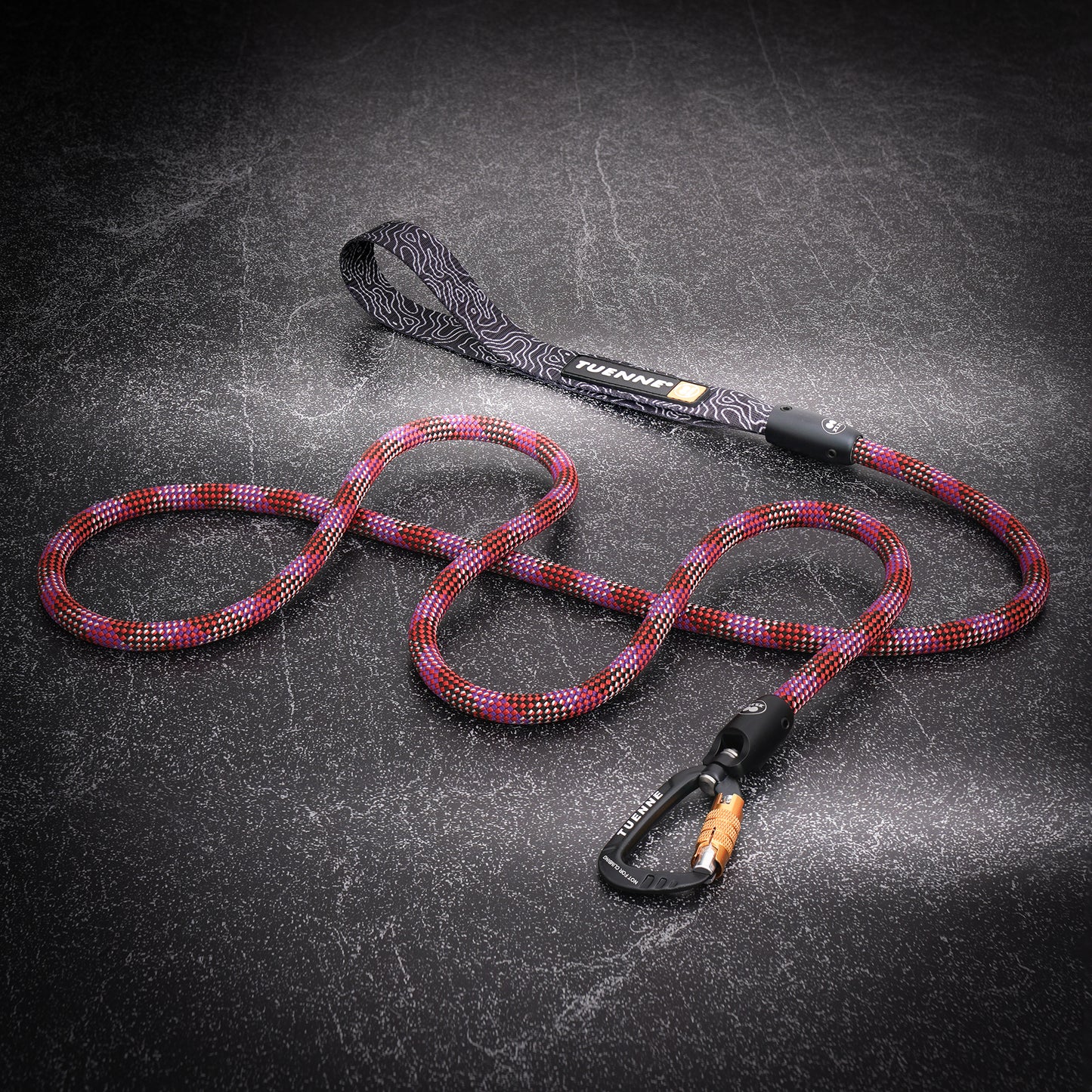 A coiled multicolor braided rope leash with a black patterned handle and a black carabiner, displayed on a dark textured background. The brand name 'Tuenne" is visible on the handle strap.