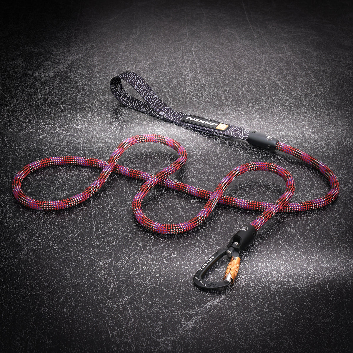 A coiled multicolor braided rope leash with a black patterned handle and a black carabiner, displayed on a dark textured background. The brand name 'Tuenne" is visible on the handle strap.