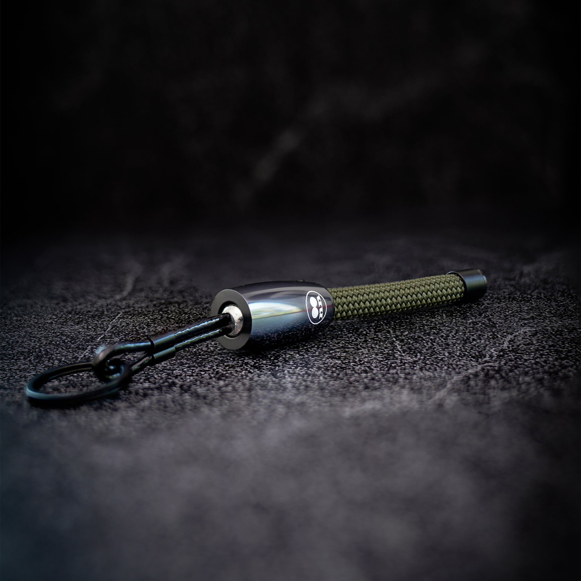 A tactical keychain or pen-style keyring with a polished silver metal body, a textured olive green rope, and a black key ring, resting on a dark textured surface.